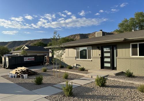 Exterior view of a contemporary home in Cedar City, UT, featuring a fresh paint job, updated modern entry door, and water-wise landscaping, with a large construction debris bin indicating ongoing work. Residential Remodel Cedar City, Utah