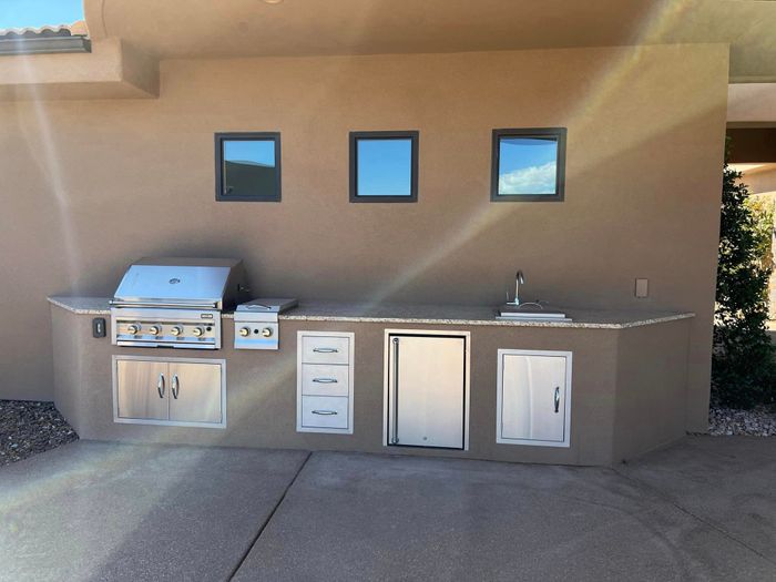 A modern outdoor kitchen with a built-in grill, stainless steel drawers, and a sink, built against a tan stucco wall with three small windows Outdoor kitchen design