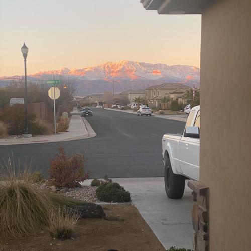 Scenic view of a quiet residential neighborhood street in Southern Utah at sunset, featuring desert landscaping, a white pickup truck in a concrete driveway, and snow-capped mountains glowing in the background. Beaitful St. George!