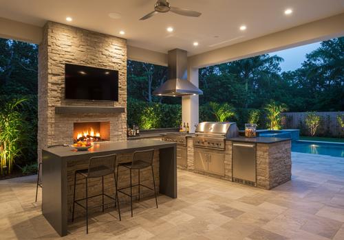 Modern outdoor kitchen and dining area featuring a built-in grill with stainless steel appliances. This luxury space includes a concrete countertop, wood cabinetry, and a large kitchen island with bar seating. The sleek, contemporary design is perfect for outdoor entertaining and al fresco dining. Keywords: outdoor kitchen, modern design, al fresco dining, grill station, kitchen island, outdoor appliances, concrete countertops, wood cabinets, backyard entertaining, luxury outdoor living. custom outdoor kitchen