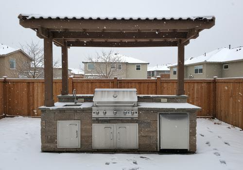 Built-in outdoor kitchen under a wooden pergola on a snowy day, highlighting the need for winterization to protect stainless steel appliances and plumbing lines. Winterized Outdoor Kitchen in St. George