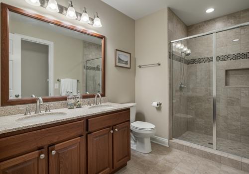 An interior view of a renovated, beige-toned bathroom. The space features a cherry wood double vanity with a speckled granite countertop and two sinks. To the right is a frameless glass walk-in shower with large beige wall tiles and a horizontal black and gray mosaic accent band. The floor is covered in matching large-format beige tiles. Completed Bathroom Renovation with Wood Vanity and Tiled Walk-In Shower.