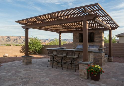 A built-in outdoor kitchen and bar with a stone base, grill, and stainless steel cabinets under a dark pergola in a backyard in St. George, Utah. St. George Outdoor Pergola and Bar