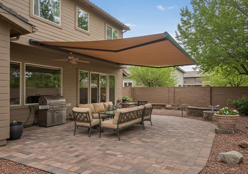 Full view of a home's backyard patio featuring a large, tan retractable fabric awning providing shade over a cushioned outdoor sofa and a stainless steel gas grill. The ground is finished with interlocking brick pavers, and the area is surrounded by a block fence. Retractable Patio Awning for Outdoor Shade
