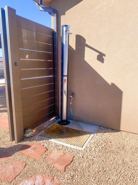 Sleek modern outdoor poolside shower fixture installed next to a dark brown slatted privacy screen on a concrete pad with gravel landscaping and red sandstone pavers. Outdoor shower