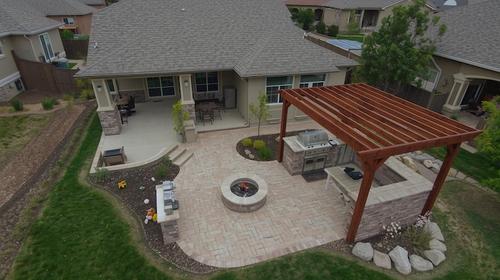 Aerial view of a full outdoor living installation, including a grill station under a wood pergola, a seating area, and a fire pit, all built with interlocking light-colored pavers extending from a home's covered patio. Aerial View of Complete Backyard Kitchen Design