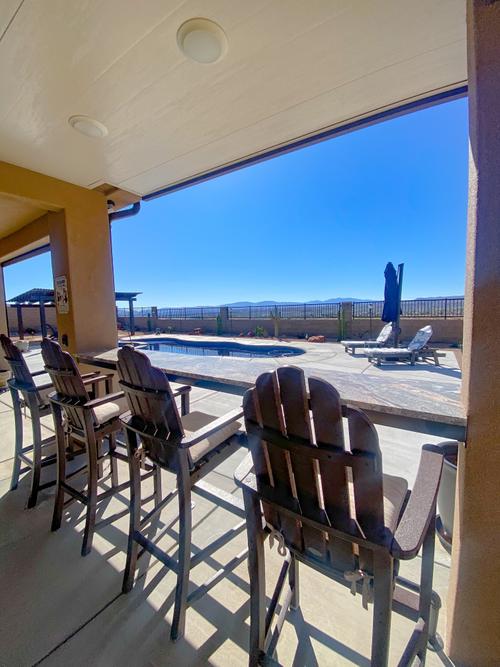 Luxury covered patio remodel featuring an outdoor bar counter with dark wood high-back swivel chairs, looking out toward a backyard swimming pool, lounge chairs, and an expansive desert horizon under a clear blue sky. An elevated outdoor living space with a poolside patio, bar seating, and mountain views in Southern Utah.