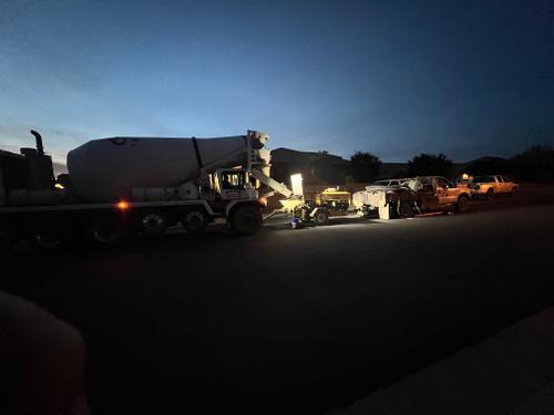 Commercial concrete mixer truck and pumping equipment set up on a residential asphalt street at night or early dawn, illuminated by bright work lights for an active construction pour. 'Crete Pouring, at Night!