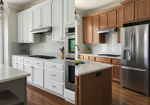 Two identical kitchen layouts shown side-by-side. The left image has white upper and lower cabinets and white countertops. The right image has medium-brown wood upper and lower cabinets and a brown wood island. Both images feature stainless steel appliances, a gray subway tile backsplash, and dark hardwood flooring. The contrast illustrates the difference in visual warmth and brightness between the two color schemes. Kitchen Cabinets: White vs. Medium Wood Tone Split Image