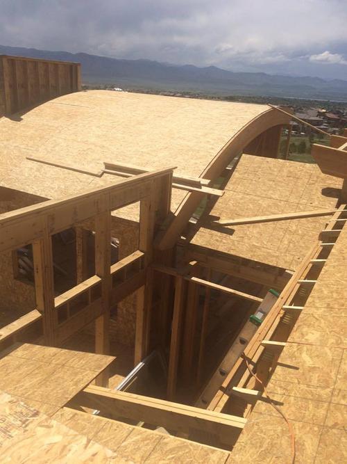 New home construction roof with a custom arched wood truss and a flat roof section. High-angle view of a custom luxury home under construction, showcasing complex curved roof framing and plywood sheathing decking on an active framing jobsite overlooking a valley.