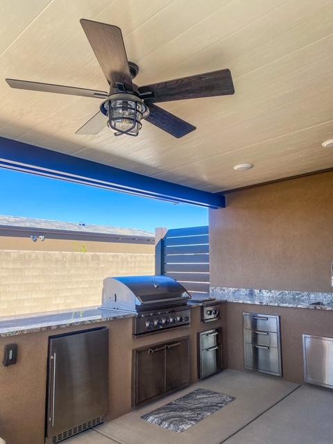 Custom covered outdoor kitchen featuring a built-in stainless steel grill, mini fridge, and storage drawers set in a stucco barbecue island with granite countertops beneath a wood paneled ceiling with a ceiling fan. custom covered patio in st george