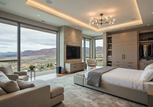 A luxurious, modern primary bedroom with floor-to-ceiling windows offering a panoramic view of red rock desert mountains and canyons. The room features a neutral color palette, a king-sized bed with a light gray upholstered frame and bench, built-in light wood wardrobes, and a television mounted on a stone accent pillar. The ceiling has recessed perimeter lighting and a modern sputnik-style chandelier. Luxury Bedroom with Desert Mountain View