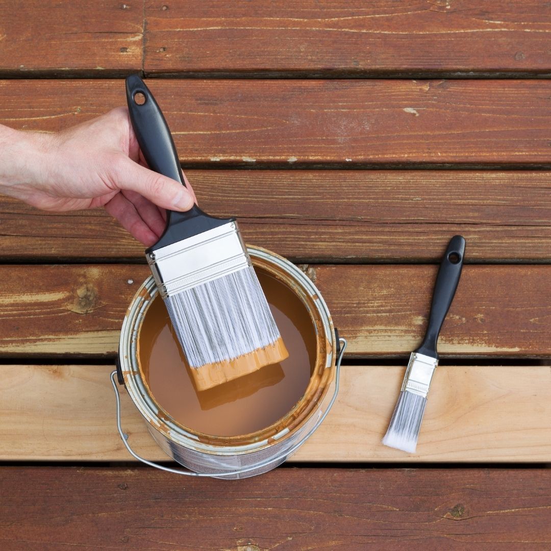 A hand dipping a brush in a can of wood stain to paint a deck