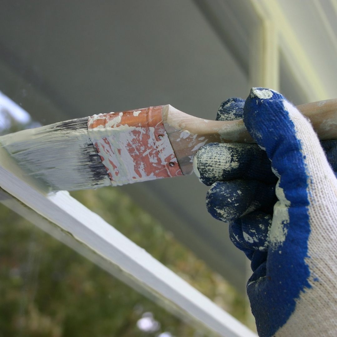 A close up of a gloved hand painting the exterior of a window's trim white