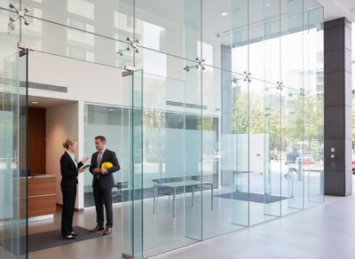 A businesswoman with a tablet talks to a businessman holding a yellow hard hat in a bright, modern office lobby with glass walls. Business professionals discussing plans in modern glass lobby