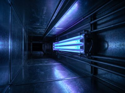 A view inside a dark, metallic commercial HVAC duct showing a glowing, vibrant blue UV light purification system. 