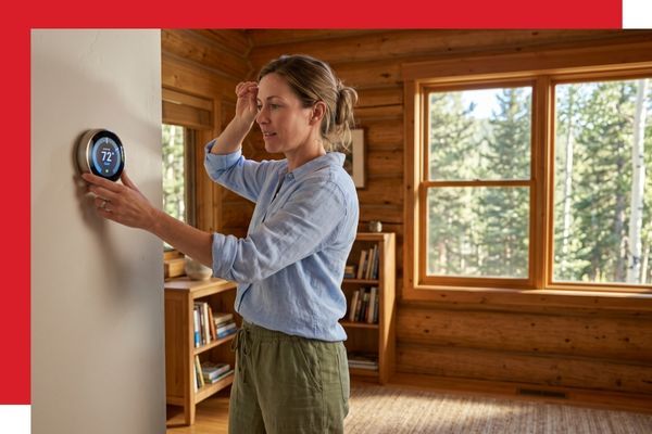 A woman in comfortable summer clothes, with hair tied up, adjusting a wall-mounted smart thermostat to lower the temperature in her rustic Allenspark, Colorado log home. A large window next to her shows a lush pine and aspen forest outside under a sunny sk