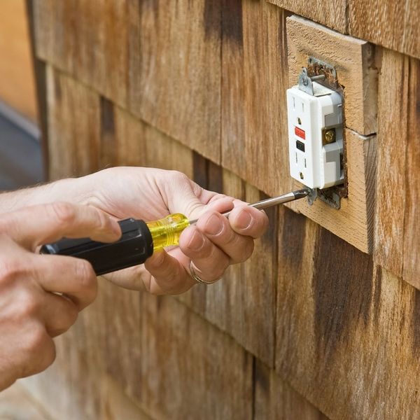 Hands of an electrician using a screwdriver to install or repair a GFI outlet on exterior wooden siding.
