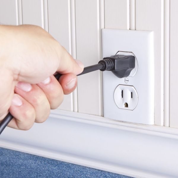 Hand plugging a black electrical cord into a white American outlet on a wainscoting wall, demonstrating correct plug insertion.