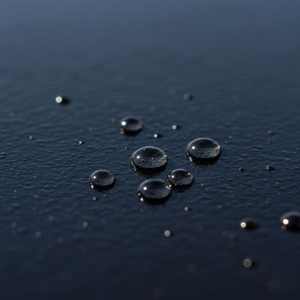 A close-up shot of a dark, freshly sealcoated asphalt surface with several small droplets of water beading up on it, showcasing its hydrophobic properties. The water droplets are distinct and round, indicating excellent water resistance. A close-up shot of a dark, freshly sealcoated asphalt surface with several small droplets of water beading up on it, showcasing its hydrophobic properties. The water droplets are distinct and round, indicating excellent water resistance.