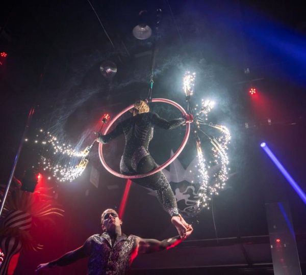 A male performer supports a female aerialist on a hoop as pyrotechnics shoot sparks into the air, creating a spectacular effect.