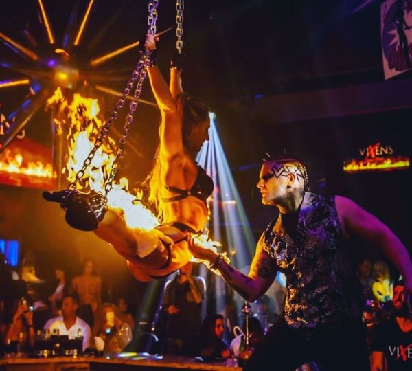 A male performer with dramatic face paint holds a flame beneath a female performer whose boots are on fire as she hangs upside down from chains in a nightclub.