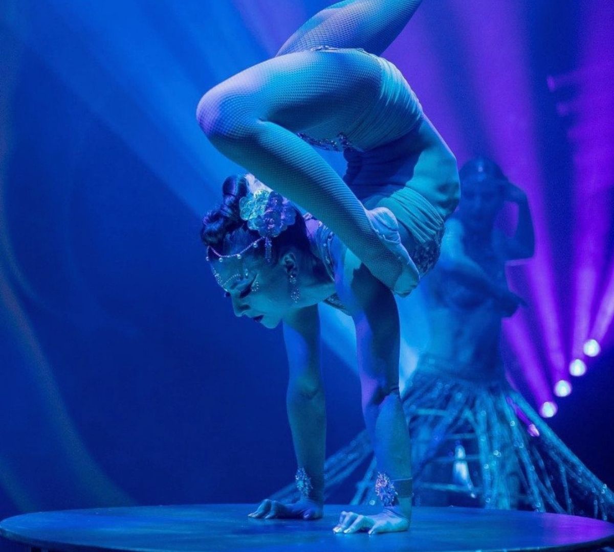 A flexible contortionist in a blue fishnet leotard performs a handstand with her back bent, illuminated by dramatic blue and purple spotlights.