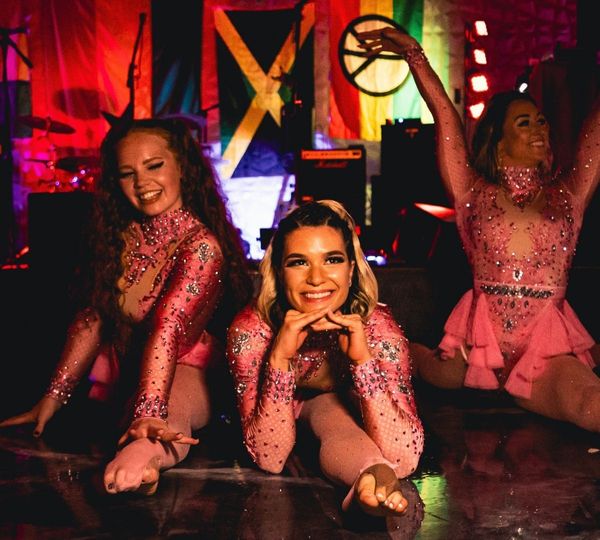 Three smiling female dancers in sparkly pink outfits pose together on a performance floor with colorful international flags behind them.