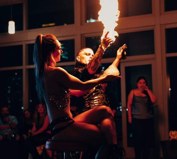A male fire performer creates a column of fire for a seated female performer in front of an audience at an indoor evening event. A male fire performer creates a column of fire for a seated female performer in front of an audience at an indoor evening event.