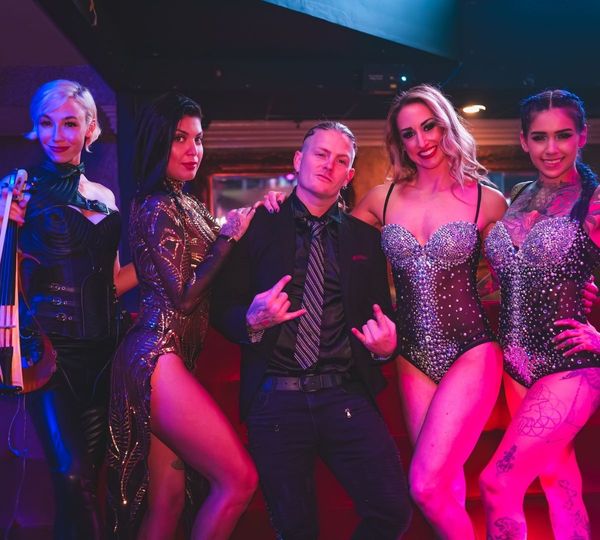 A male performer in a black suit makes a "rock on" gesture while posing with four female performers in elaborate, glittering cabaret costumes under vibrant stage lights.