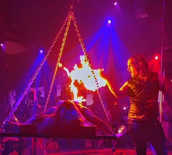 A male performer with dramatic makeup directs a large flame towards a woman lying on a table in a theatrical performance with red and blue stage lighting. A male performer with dramatic makeup directs a large flame towards a woman lying on a table in a theatrical performance with red and blue stage lighting.