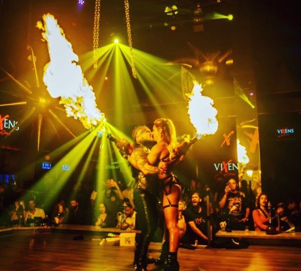 Two performers embrace in a kiss while holding large, lit, flame throwers, illuminated by bright yellow spotlights in a crowded event.