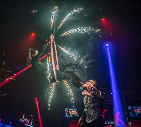 A male performer supports a female aerialist by her feet as she hangs from a strap, surrounded by a fan of glowing sparklers.