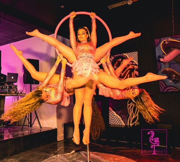 Three female aerialists in matching glittering pink costumes perform a complex, synchronized pose on a single hoop at an event. Three female aerialists in matching glittering pink costumes perform a complex, synchronized pose on a single hoop at an event.