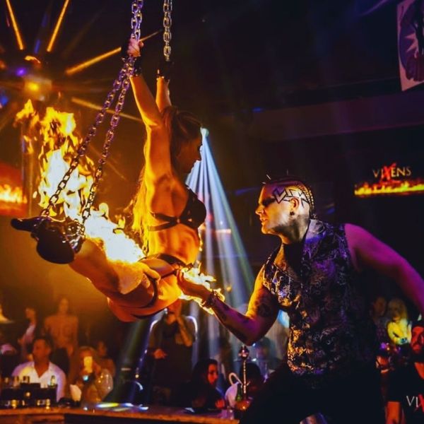 A fire performer suspended by chains over an audience, with flames coming from their feet and hands, and a male performer engaging with them in a nightclub setting.