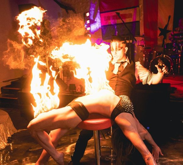 In a dramatic performance, a male fire artist holds a large flame above a female performer who is arched backwards over a stool.