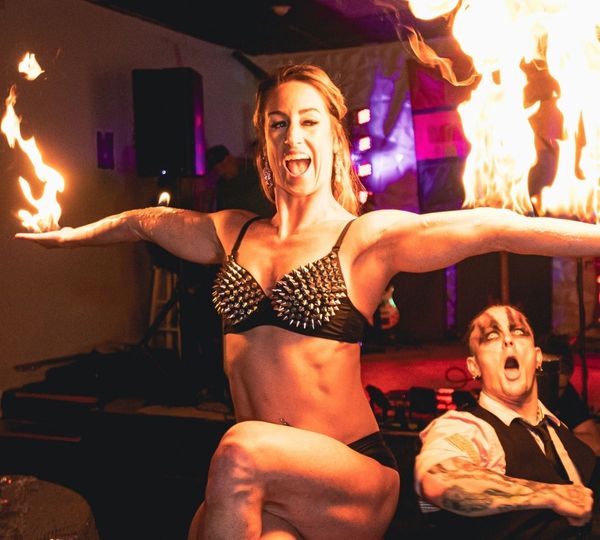 A muscular female performer in a studded bra smiles as she holds two flaming batons, while a male performer looks on with a shocked expression. A muscular female performer in a studded bra smiles as she holds two flaming batons, while a male performer looks on with a shocked expression.