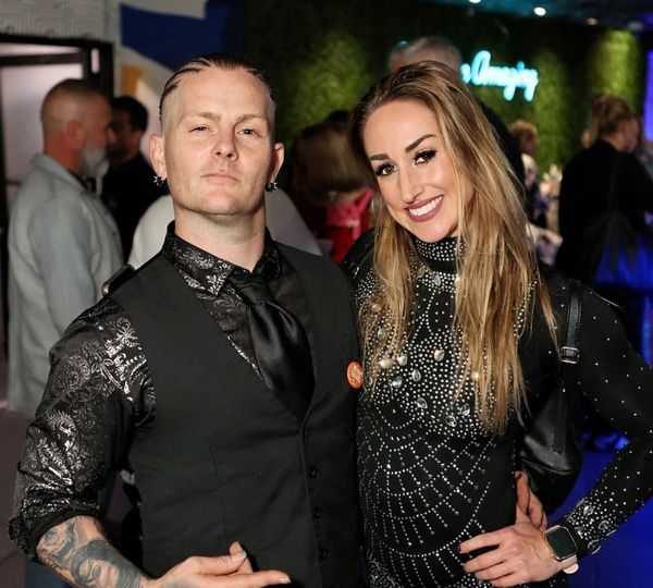 A man with tattoos and a formal vest stands with a smiling woman in a stylish, jewel-studded black dress at a corporate event.
