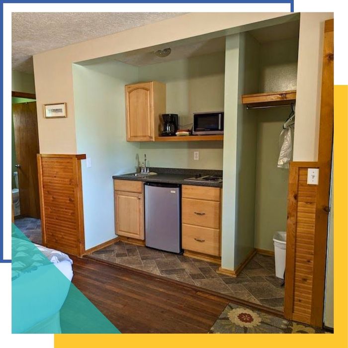 A brightly lit, functional kitchenette area inside a deluxe room at Mitchell Creek Inn, showing light wood cabinets, a sink, a two-burner cooktop, a microwave, and a mini-fridge