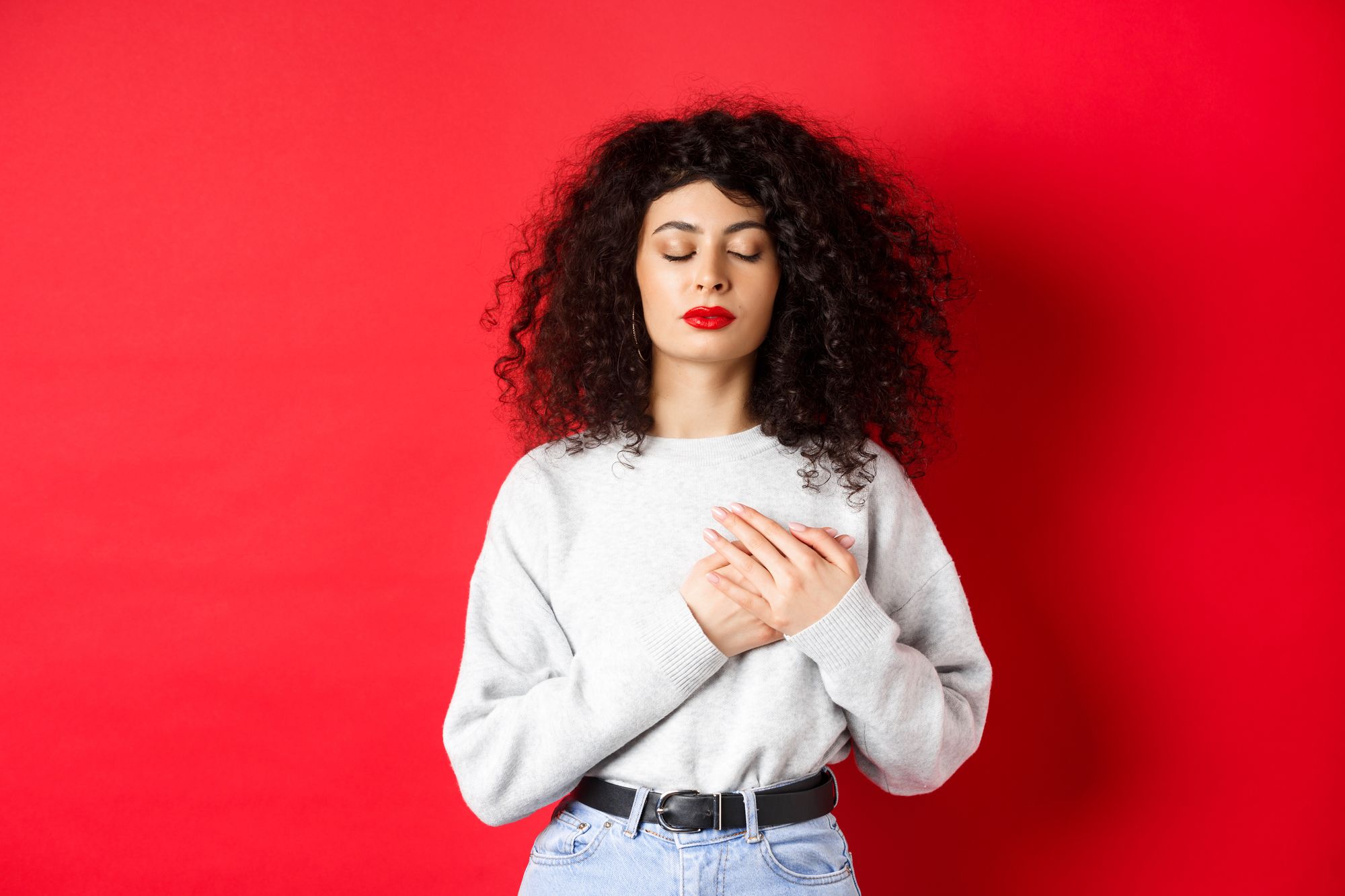 image-calm-young-woman-with-curly-hairsty-close-eyes-holding-hands-heart-keeping-warm-memories-feeling-nostalgic-standing-red-background.jpg