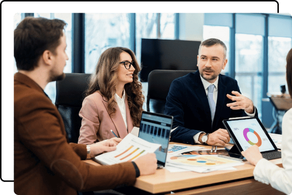 A business meeting with a man in a suit speaking to two people across a table, looking over documents and a laptop, discussing marketing strategy. A business meeting with a man in a suit speaking to two people across a table, looking over documents and a laptop, discussing marketing strategy.