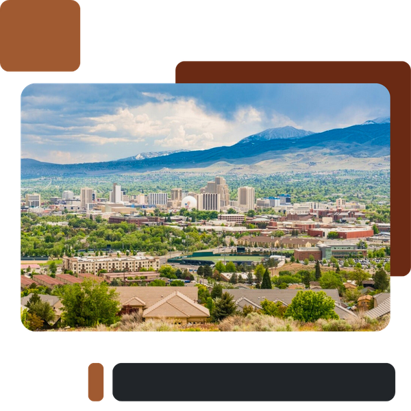 A wide panoramic view of the downtown Reno, Nevada skyline, featuring tall buildings nestled among green trees, with residential areas in the foreground and the snow-capped Sierra Nevada mountains in the background under a dramatic cloudy sky.