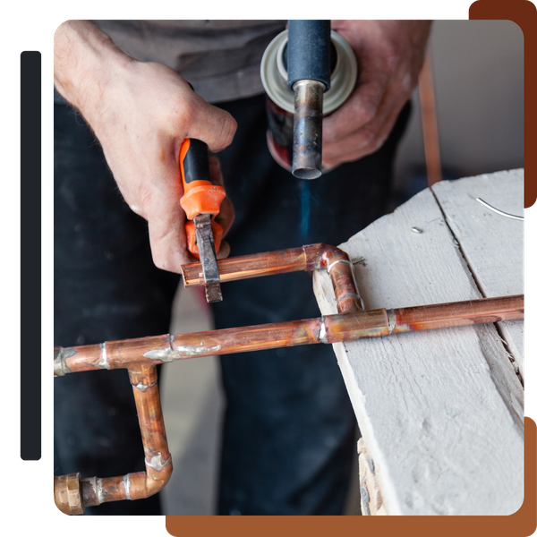 A close-up of a person's hands using a torch to heat and solder two copper pipes connected at a joint, with a pair of pliers resting nearby.