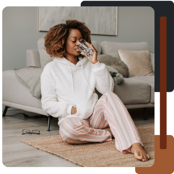 A young woman with curly hair, wearing a white fluffy sweater and striped pajamas, sitting on a rug and drinking a glass of water in a relaxed home setting.