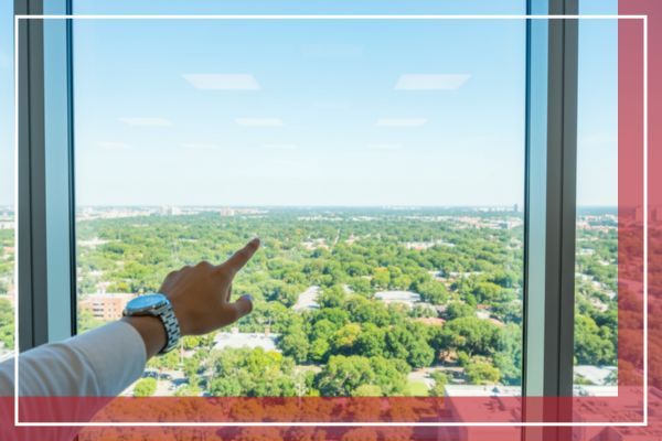 A person's hand points out a large, clear office window, showcasing an unobstructed panoramic view of a lush green cityscape in the distance on a bright day.