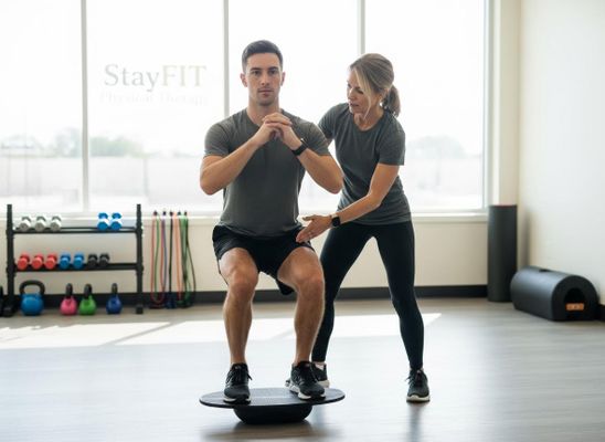 A man in a grey shirt and black shorts squats on a black balance board, supported by a female physical therapist in a gym. Man squatting on balance board with physical therapist assistance