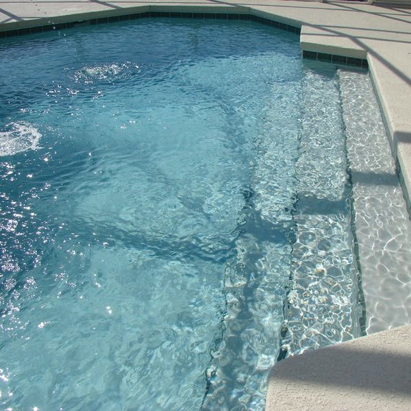 A close-up view of the clear, blue water of an inground swimming pool with wide, textured steps leading into the shallow end. Sunlight creates shimmering patterns on the water's surface. A close-up view of the clear, blue water of an inground swimming pool with wide, textured steps leading into the shallow end. Sunlight creates shimmering patterns on the water's surface.