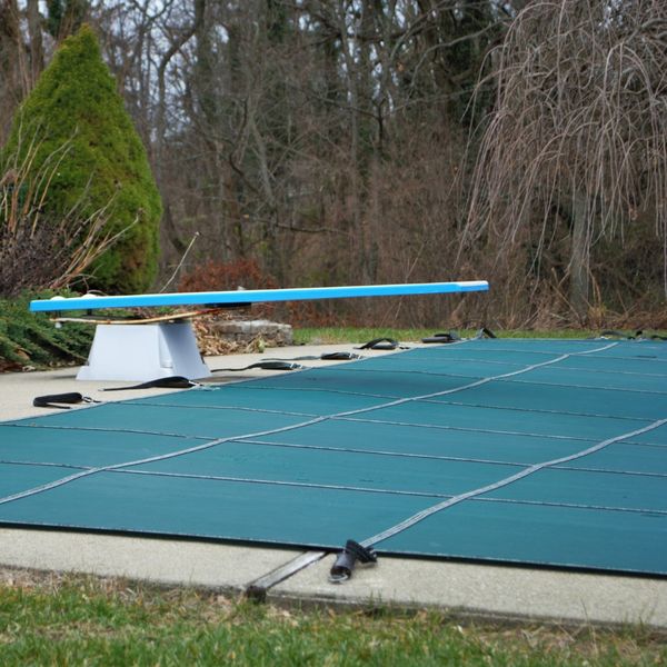 An inground swimming pool covered with a dark green winter safety cover, secured with straps. A blue diving board extends over the covered pool. Bare trees and evergreens are visible in the background, suggesting autumn or winter. An inground swimming pool covered with a dark green winter safety cover, secured with straps. A blue diving board extends over the covered pool. Bare trees and evergreens are visible in the background, suggesting autumn or winter.