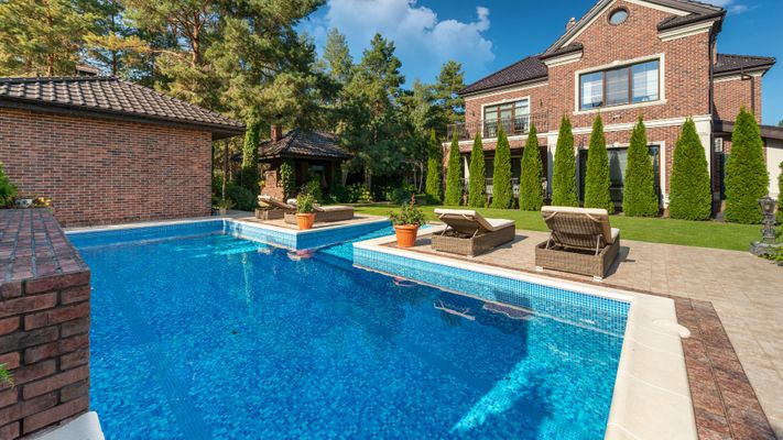 A luxurious backyard with a large, L-shaped inground swimming pool featuring blue mosaic tiles and a wide, tiled sun shelf. Two lounge chairs with orange potted plants sit on a raised section of the pool deck. In the background is a large brick house, a sm