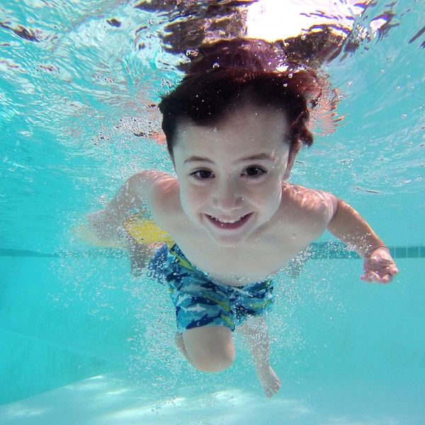A young boy with light brown hair and blue swimming trunks is underwater in a bright blue swimming pool, smiling directly at the camera with bubbles around him. A young boy with light brown hair and blue swimming trunks is underwater in a bright blue swimming pool, smiling directly at the camera with bubbles around him.
